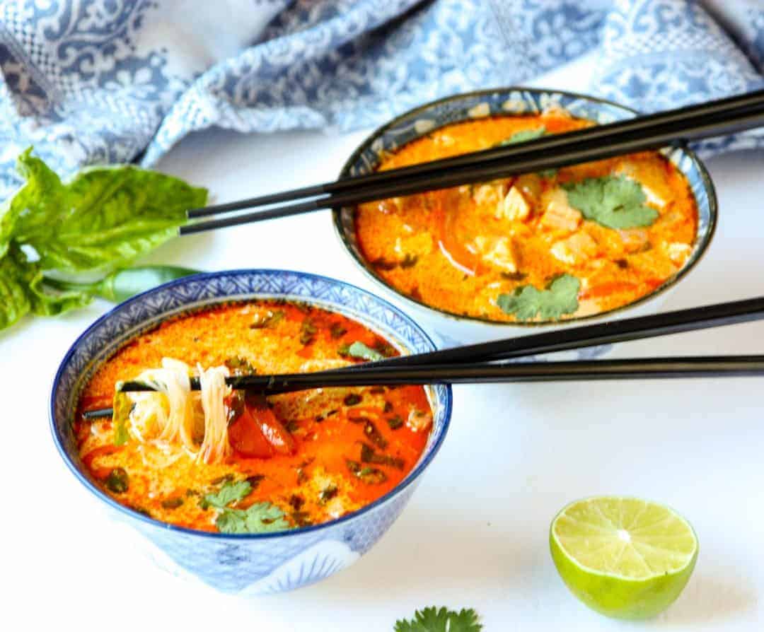 Two blue & white patterned bowls of red thai curry chicken soup with noodles next to a sprig of basil and wedge of lime